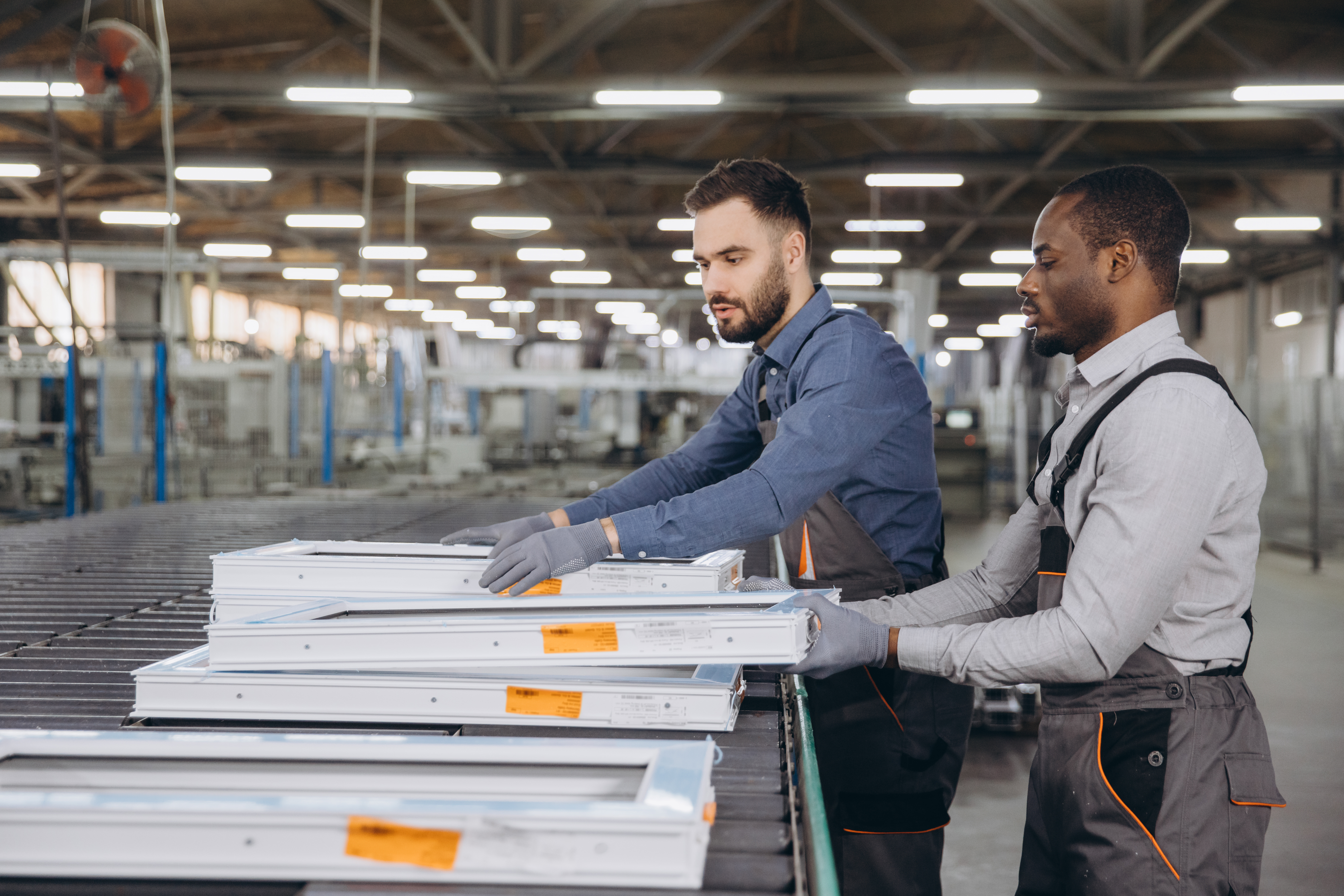 Two men in work uniforms and gloves inspect and stack metal window frames on a conveyor belt, demonstrating fabrication efficiency inside a large, well-lit factory that supports operations at a UK nuclear site.
