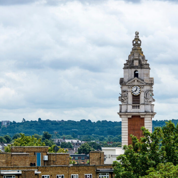 A tall clock tower with a brick base and ornate top stands among trees and ModernaHus residential scheme buildings, with a cloudy sky and distant green hills in the background.