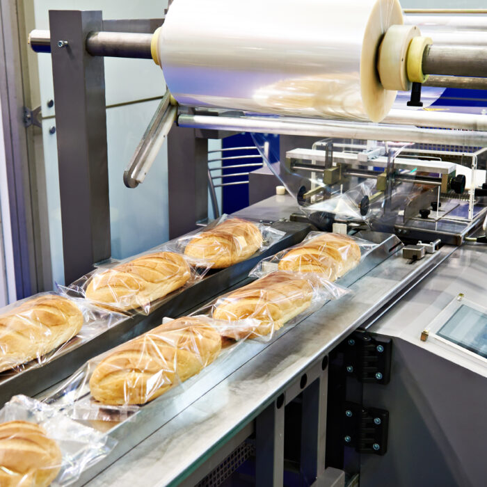 At a Premier Foods facility, a commercial bread packaging machine wraps loaves in clear plastic film; several packaged loaves move along a conveyor belt, with the control panel and roll of plastic visible—ideal for food manufacturing operations.