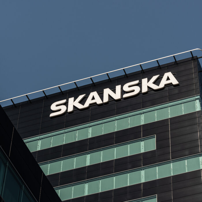 A modern office building with large windows and "Skanska" in bold white letters near the top, set against a clear blue sky, adjacent to a new build workshop facility.