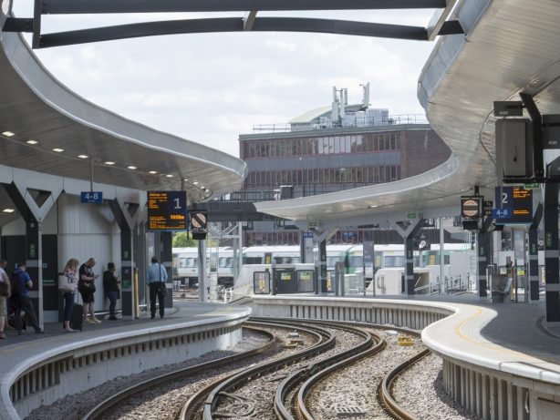 Curved train tracks run through the modern London Bridge Station, where several platforms host waiting passengers by digital display boards. A train is visible in the background near a large building under a partly cloudy sky.