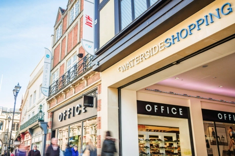 People walk past the Waterside Shopping Centre, with visible OFFICE and New Look store signs on a busy street lined with shops and historic-style buildings in the heart of the city.
