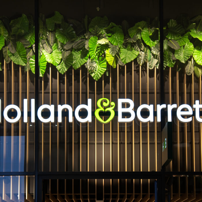 A brightly lit “Holland & Barrett” store sign with a green ampersand, displayed under a row of large green leaves, hints at the food warehouse redevelopment transforming the storefront’s appearance.