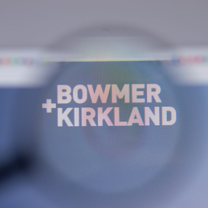 A computer screen shows the Bowmer & Kirkland logo, viewed through a magnifying glass, with the company name in bold white text on a blurred background—highlighting their work on Liverpool's Kings Dock Mill Residential Scheme.