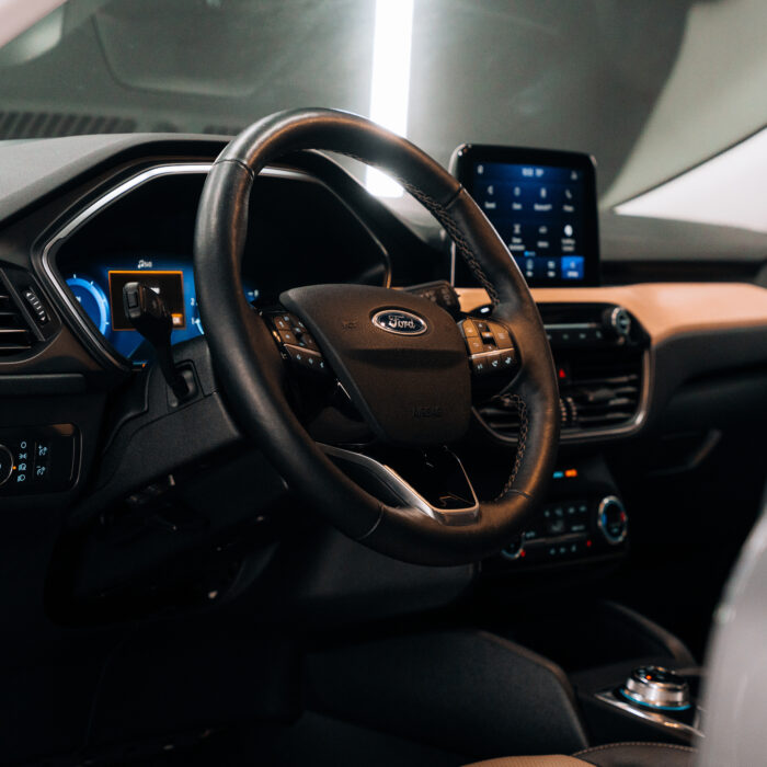 Close-up view of a modern car interior featuring a black Ford steering wheel, digital dashboard, and Project DV Neo touchscreen display, with various control buttons on the dashboard and center console illuminated by ambient lighting.