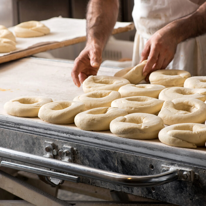 A baker shapes rings of raw dough into bagels on a metal worktable, with trays of unbaked bagels nearby in a busy New York Bakery setting.
