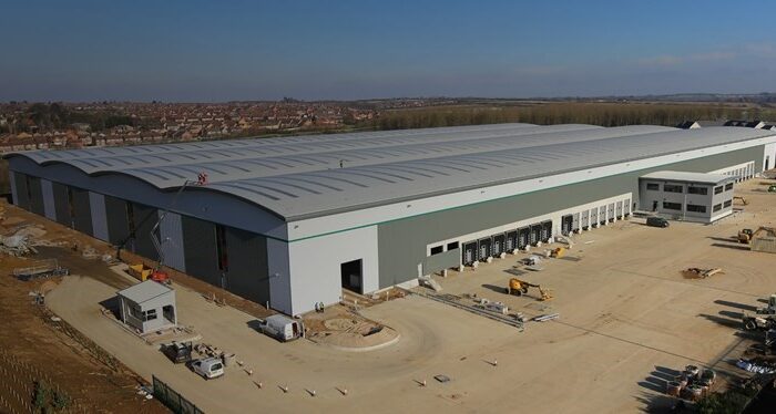 Aerial view of a large, modern distribution centre under construction in Grange Park, surrounded by equipment, vehicles, and cleared land, with a suburban neighborhood visible in the background.