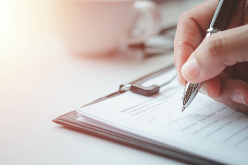 Close-up of a hand holding a pen and filling out a Building Inspection form on a clipboard, with a blurry coffee cup in the background.