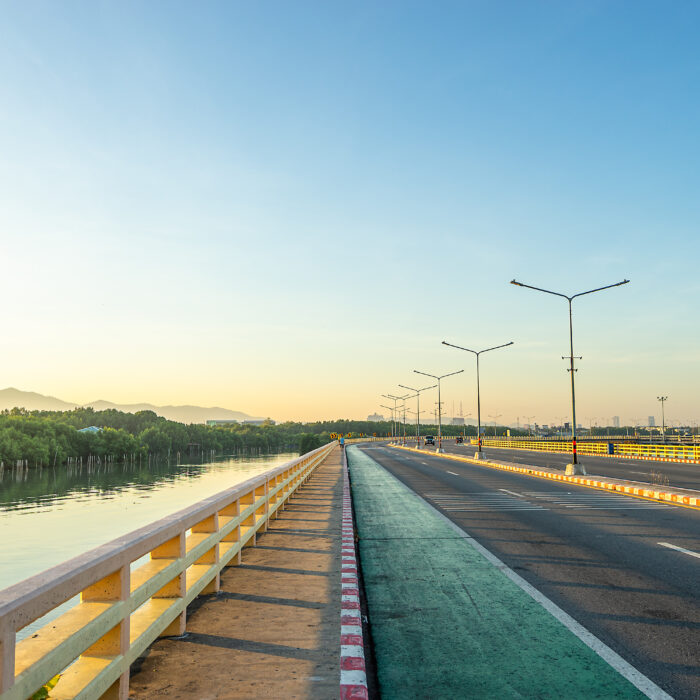 A wide, empty road with streetlights runs alongside a river, bordered by yellow railings, under a clear blue sky at sunrise—an ideal scene for Strategic BIM to support infrastructure planning amid distant mountains and lush greenery.