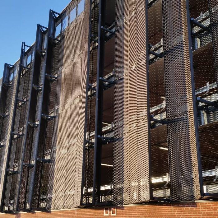 The Lincoln Transport Hub features a modern building with a red brick base and large vertical metal mesh panels covering multiple stories, creating a geometric pattern, seen in bright sunlight under a clear blue sky.