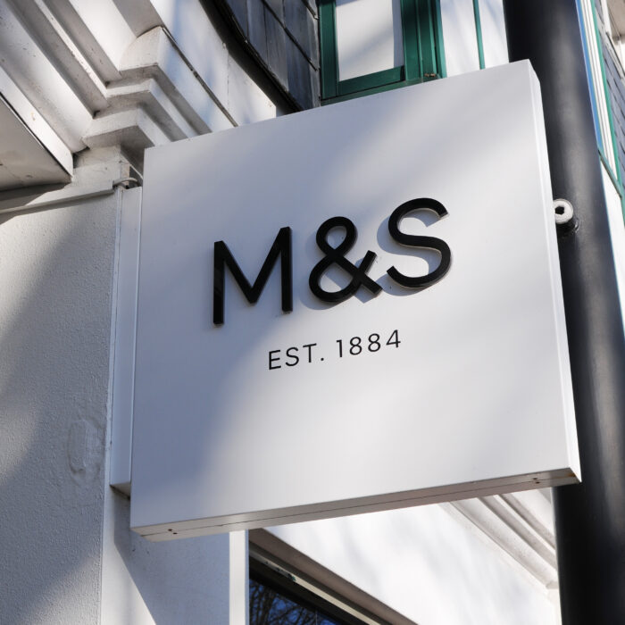 A white square sign with a concept design is attached to an inner town building, displaying the black letters "M&S" and the text "EST. 1884" below it.