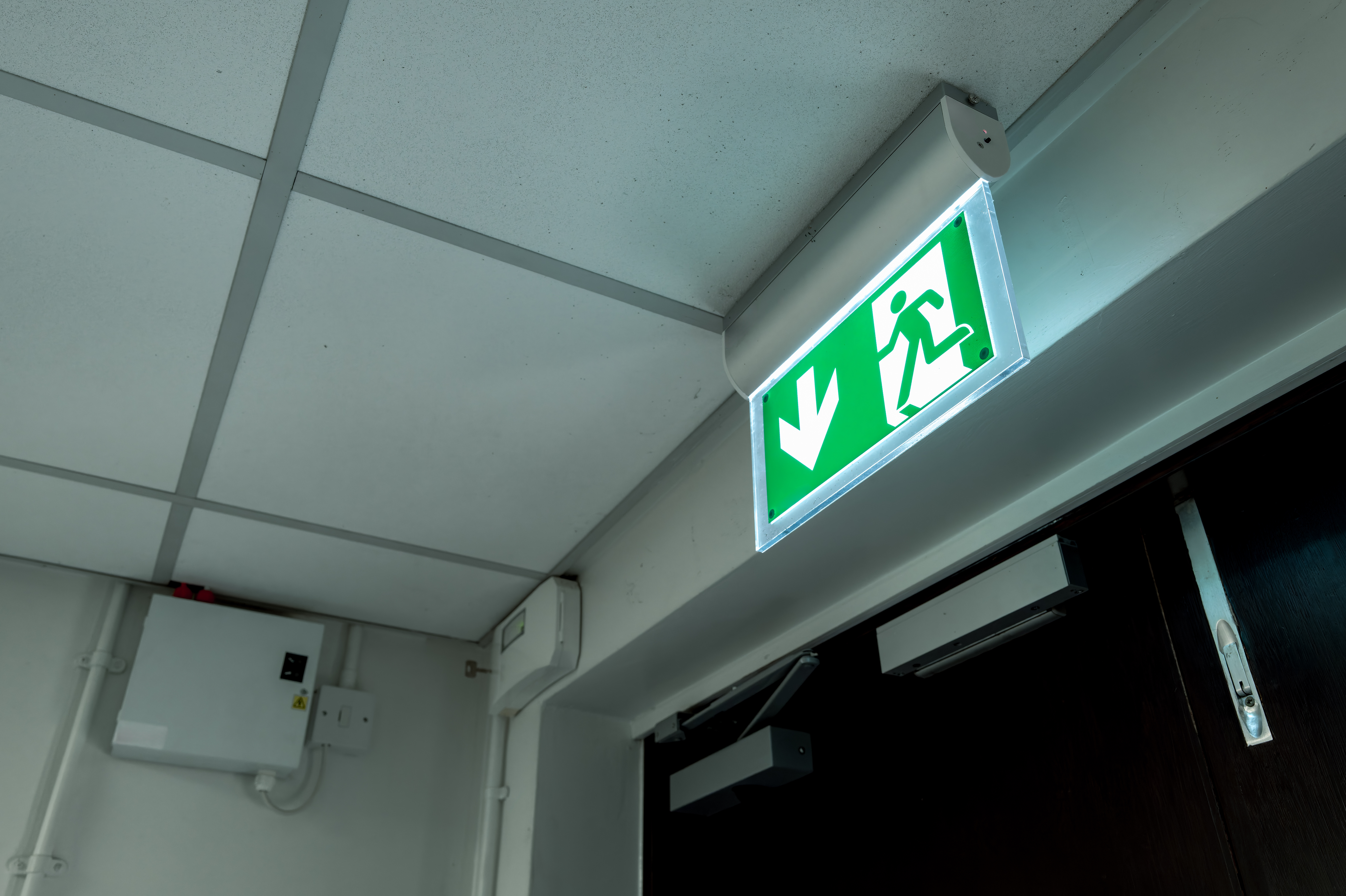 A green emergency exit sign with a running figure and downward arrow is illuminated above a doorway in an indoor setting at HMYOI Deerbolt, reflecting ongoing Fire Safety improvements amidst white ceiling tiles and visible electrical wiring.