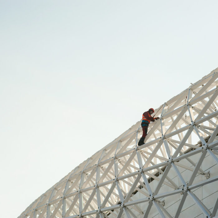 A worker in safety gear climbs the metal framework of a large structure at HMP Warren Hill, where refurbishment works are underway, set against a bright, clear sky.