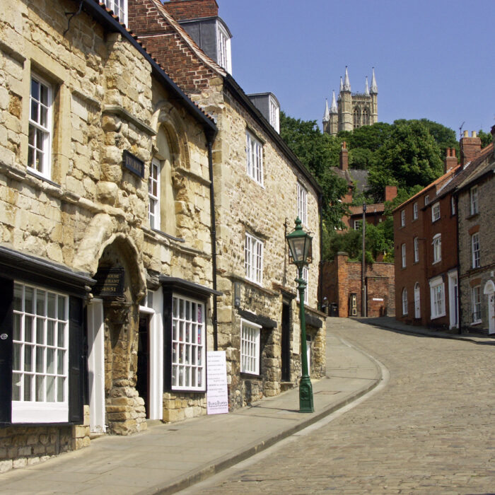 A cobblestone street lined with old stone buildings, some undergoing refurbishment, leads uphill toward large cathedral towers, partially hidden by trees, under a clear blue sky.