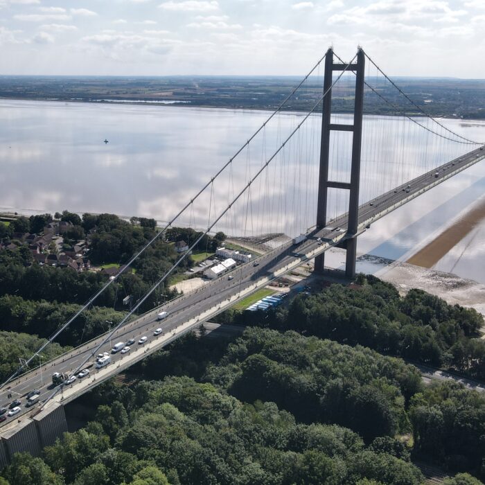 A long suspension bridge spans a wide river, connecting two distant shores. Cars cross the bridge, while green trees and houses line one side and open water reflects the sky—just beyond, HMP Humber’s Control Room Migration is underway in the background.