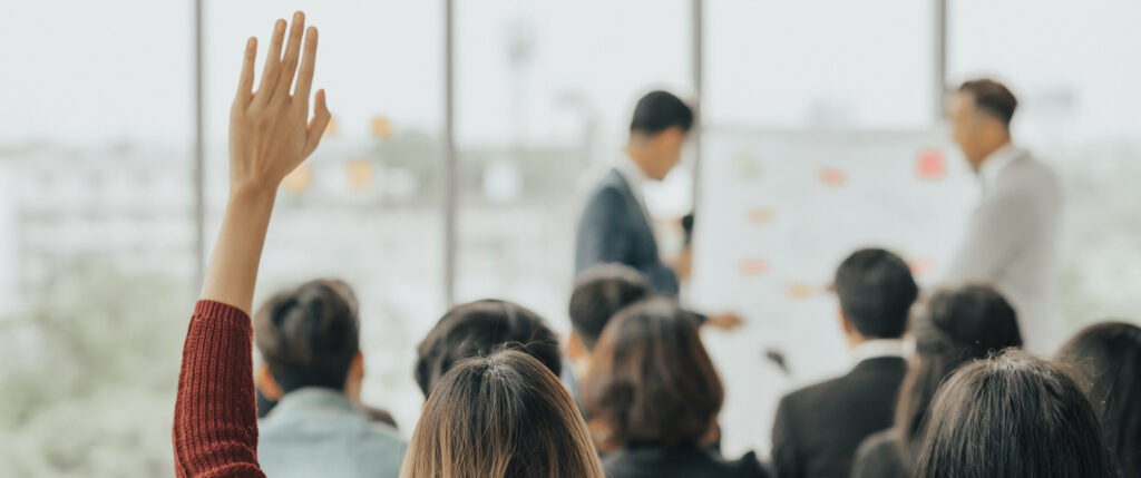 A person raises their hand in an audience while two speakers present information about the ICE approved training scheme at the front of a bright, modern room with large windows.