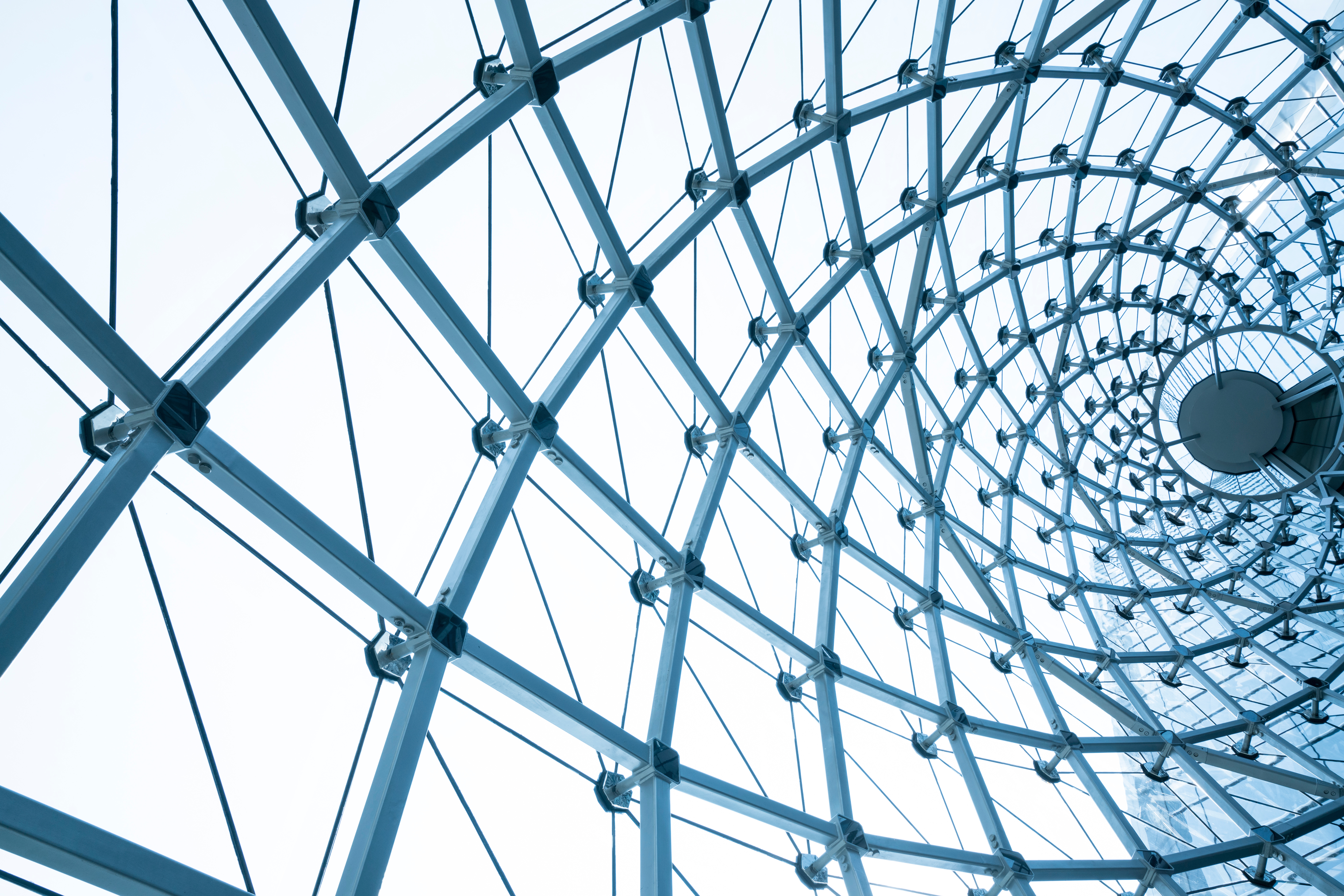 View looking up at a modern glass and steel dome ceiling with a spiral pattern, showcasing geometric symmetry against a bright sky—a stunning example of architecture found in Secure Place and projects led by Waldeck.