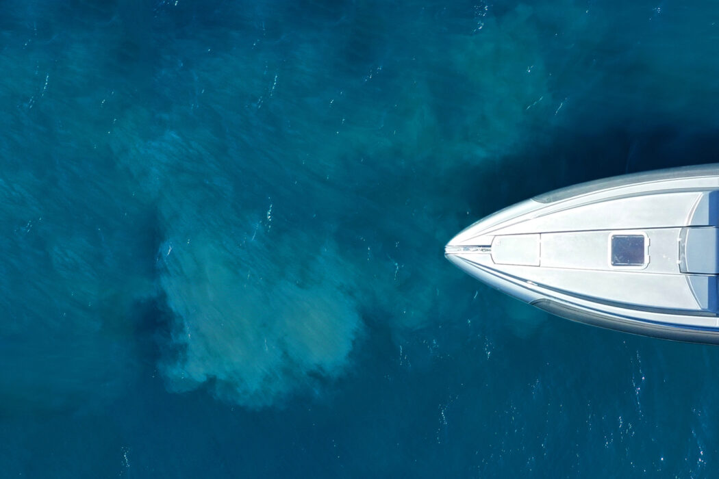 Aerial digital capture of a luxury yacht floating on calm, clear blue water, with the boat positioned on the right and deep, vibrant ocean visible on the left.