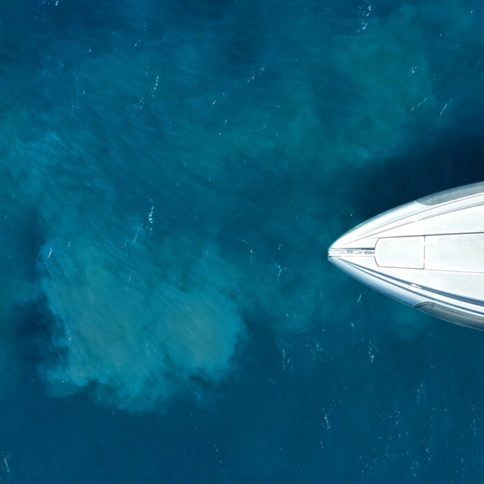 Aerial digital capture of a luxury yacht floating on calm, clear blue water, with the boat positioned on the right and deep, vibrant ocean visible on the left.