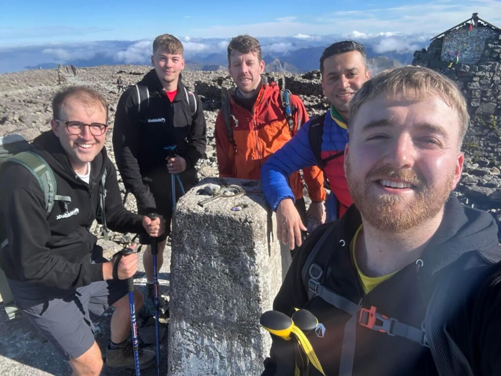 Five men in hiking gear smile for a selfie at the summit, gathered around a white concrete marker. Civil engineer Thomas Peden, known as a mentor to many, stands with them under sunny blue skies and distant mountains.