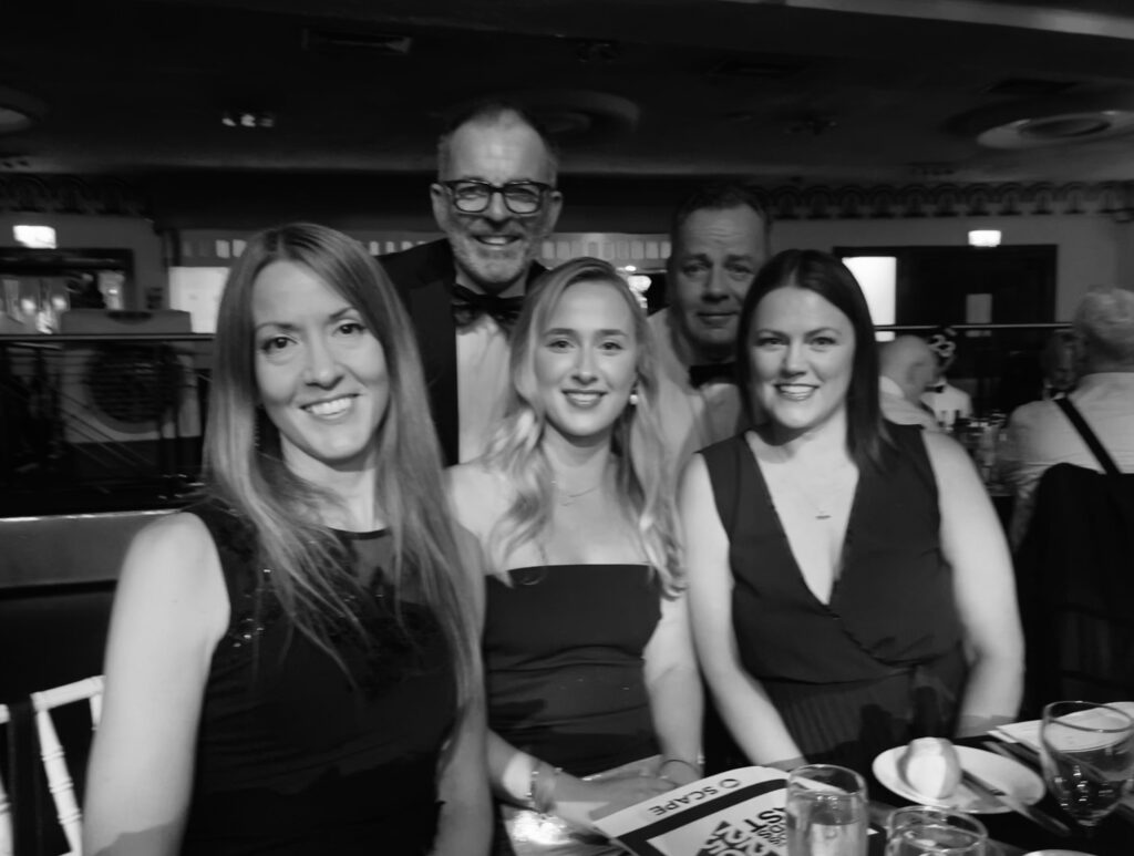 Five adults, including Nina Windmann, Marketing & Communications Assistant, pose at a formal indoor event. Three women are seated in front with two men standing behind; all smile amid dinner tables and elegant decor. The photo is in black and white.