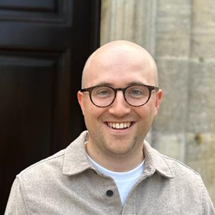 Thomas Peden, a smiling civil engineer and mentor with glasses and a bald head, stands outside in front of a stone wall and a dark door, wearing a light beige collared shirt.