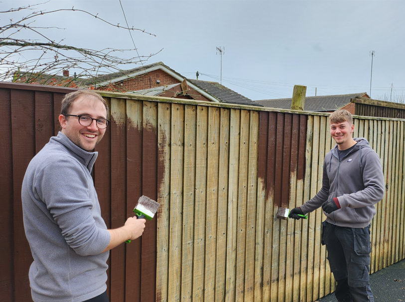 Two people, including mentor and civil engineer Thomas Peden, smile while painting a wooden fence outside. One section is brown and finished; the other is still bare wood. Both hold paintbrushes and wear casual outdoor clothing.