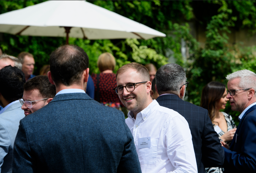 A group of people are socializing outdoors under a white umbrella. Civil engineer Thomas Peden, in a white shirt and glasses, smiles while talking to another man in a blue jacket. Lush greenery is visible in the background.