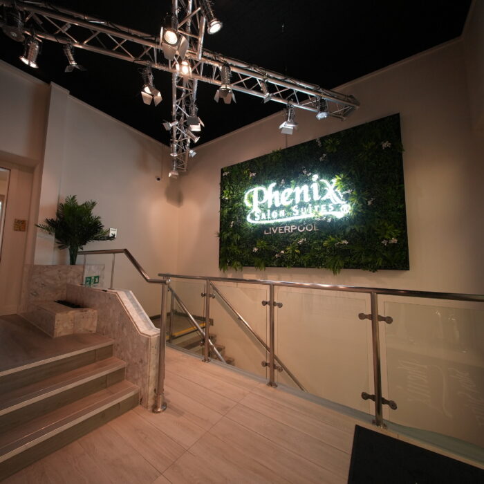 A modern interior with a lit sign reading "Phenix Suites LIVERPOOL" on a plant-covered wall, showcasing stylish Liverpool Salon Units, stairs with a marble railing, glass balustrade, and ceiling spotlights.
