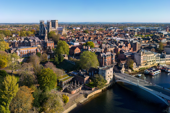 Aerial view of York, England, showing the historic York Minster cathedral, the River Ouse, Lendal Bridge renowned for its bridge engineering, and a mix of old buildings and greenery under a clear blue sky.