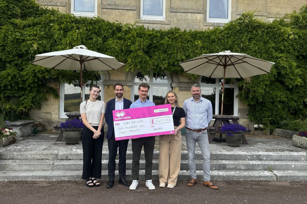 Five people stand outdoors in front of a building with greenery, holding a £640 ceremonial check for Cash for Kids. The Waldeck Wellingore Team presents the donation, with two large umbrellas visible behind them.