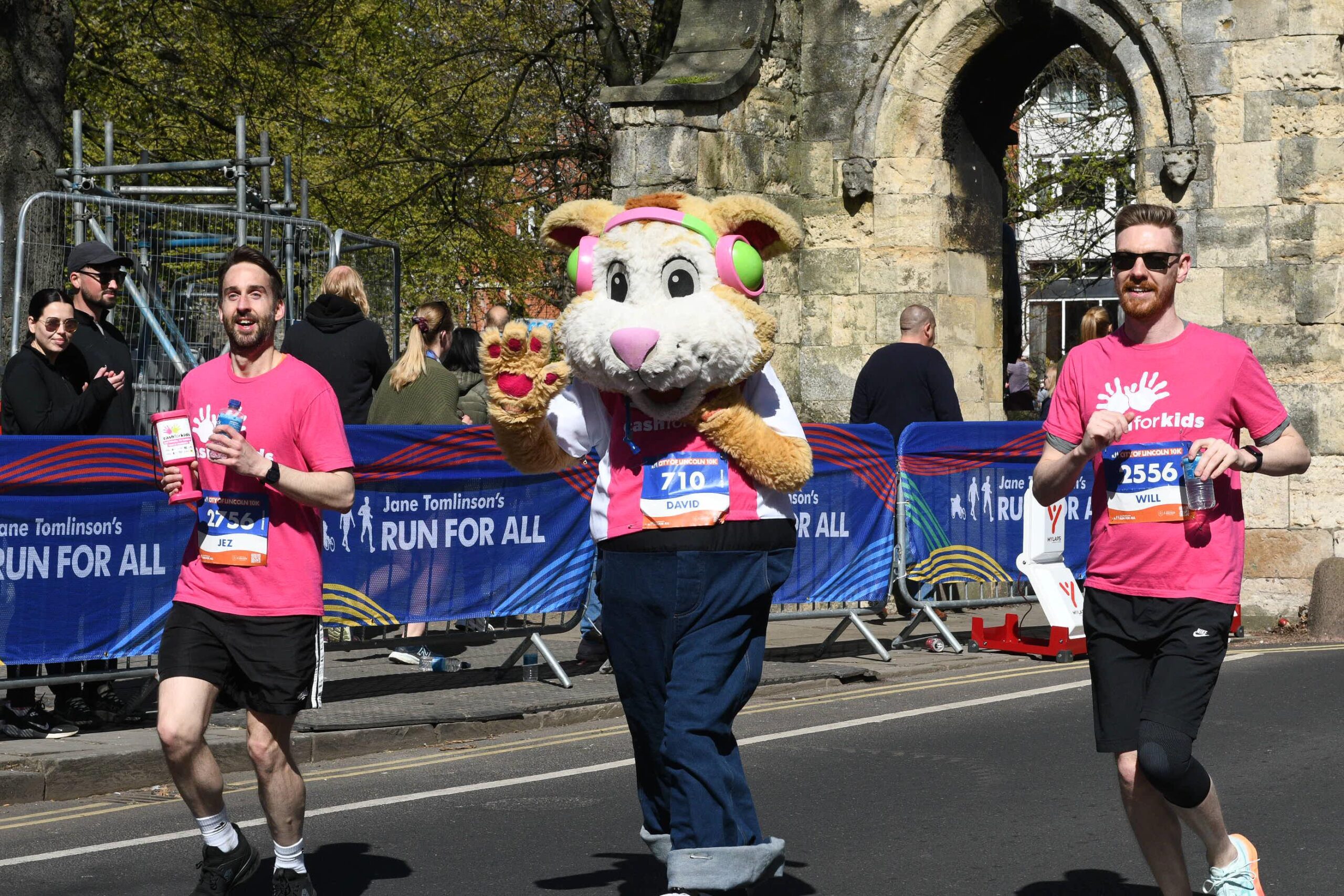 Three runners participate in a race; two men in pink shirts run alongside a person in a large animal mascot costume. They smile and wave near a stone archway and blue race banners.