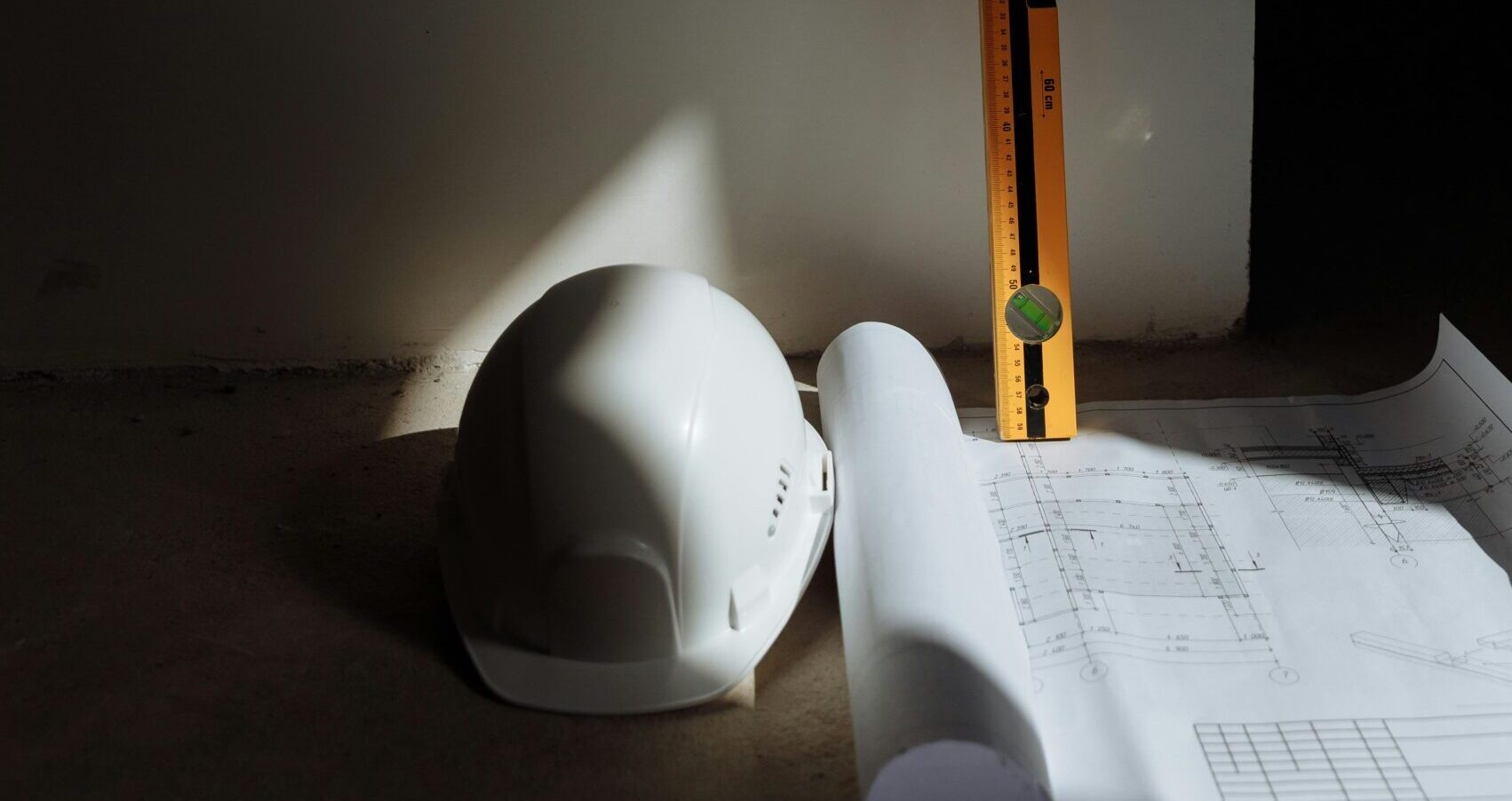 A white construction helmet, a rolled-up blueprint, and a yellow spirit level are placed on the floor against a plain wall, illuminated by a shaft of light—symbolizing progress towards Net Zero Goals in construction.