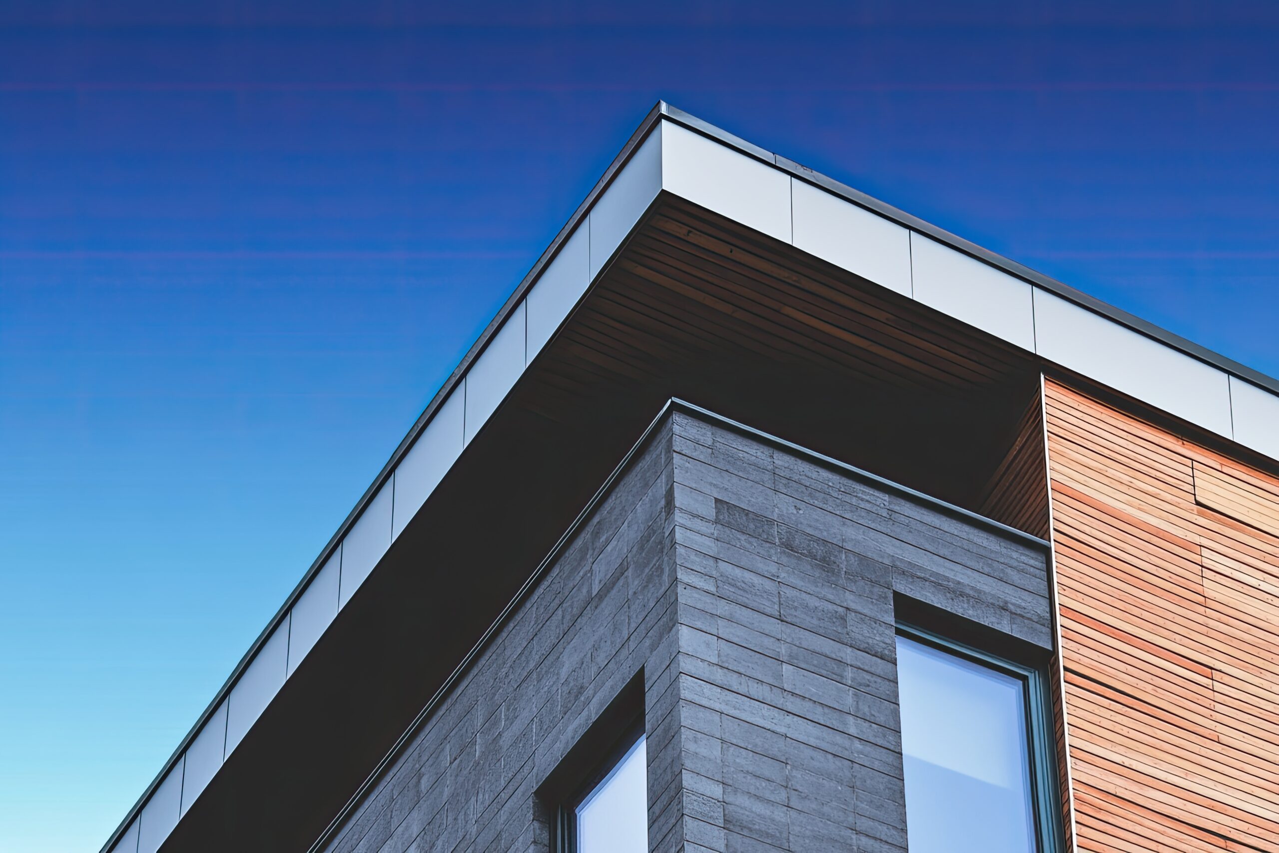 Close-up view of a modern building corner by Waldeck, featuring gray concrete, wood paneling, large windows, and a clear blue sky—delivered under the EN:Procure Professional Services Framework.