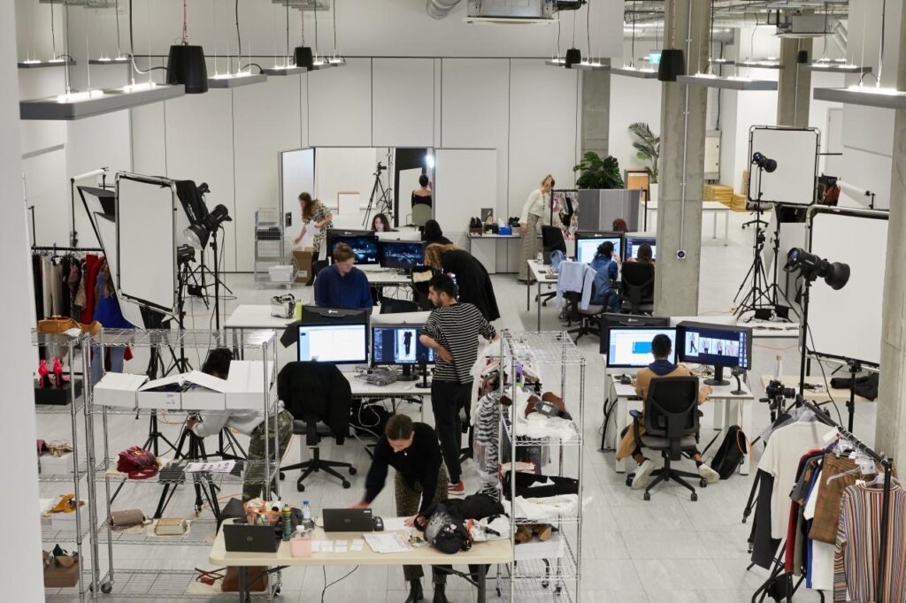 A modern photo studio with people working at desks and computers. Photography equipment, lighting, and clothing racks are visible, while teams edit images and arrange clothes in an open, well-lit workspace that promotes employee wellbeing.