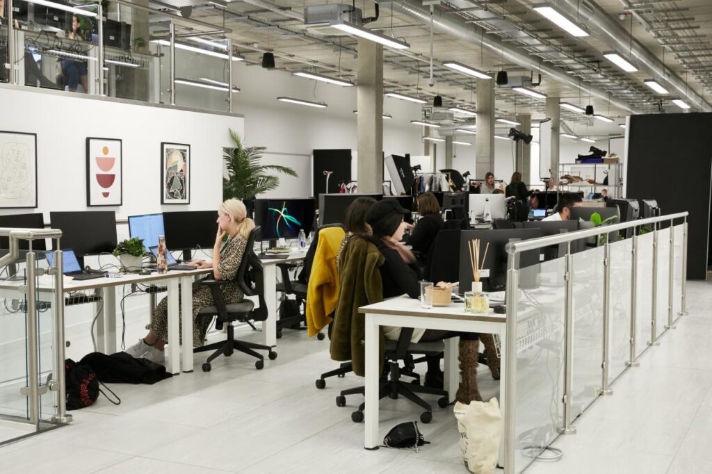 Modern open-plan office with people working at desks on computers. The space, designed with employee wellbeing in mind, features white walls, exposed ceilings, and contemporary décor that fosters collaboration and healthier workplaces.