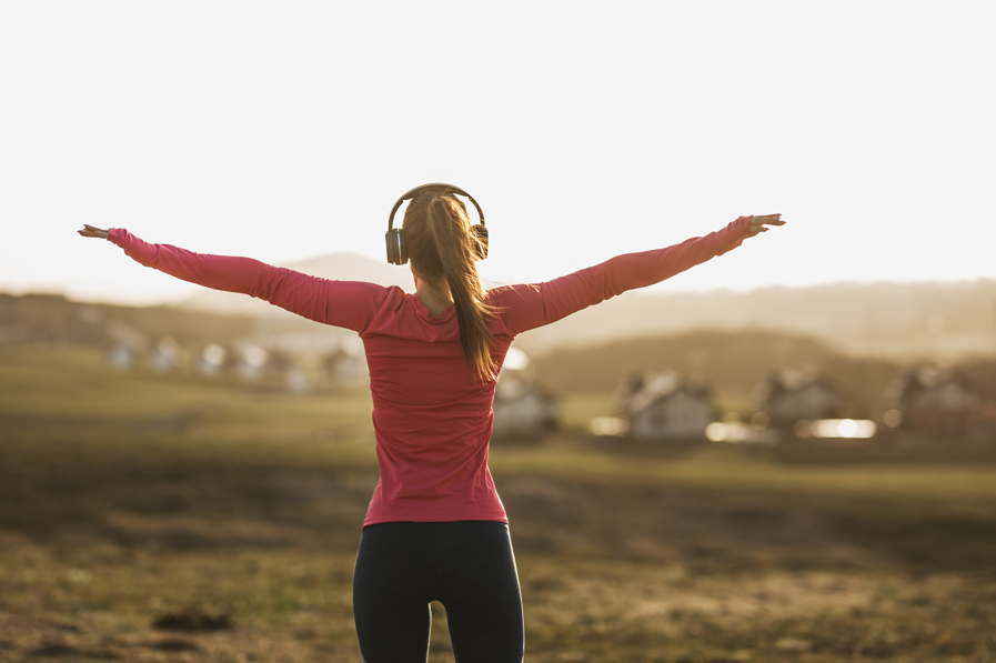 A person embodying workplace wellbeing stands outdoors in headphones, a pink long-sleeve shirt, and dark leggings, arms outstretched toward a blurred landscape with houses and fields at sunset.