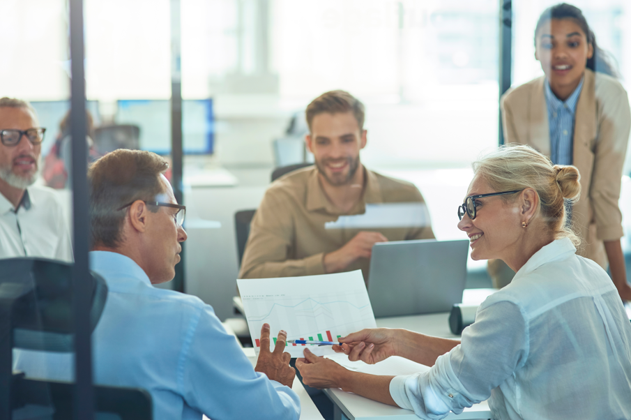 Five colleagues in business attire meet in a bright office. A woman in glasses presents a graph to a man, while three others look on and smile, discussing Workplace Wellbeing. Laptops and papers are on the table.