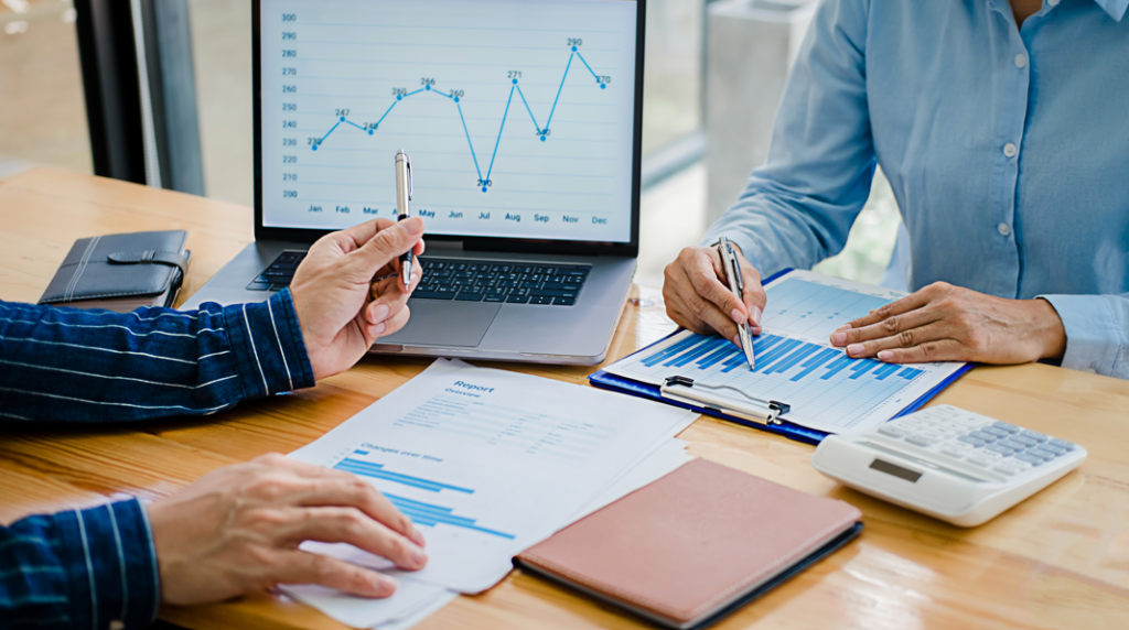 Two people review financial charts and graphs at a desk, aiming to enhance workplace wellbeing. Papers, a laptop with a line graph, a clipboard, notebook, and calculator are spread out as they discuss key building consultancy services.