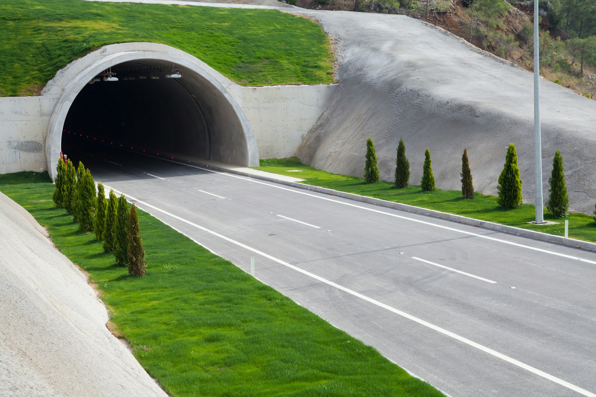 A wide, paved road leads into a tunnel entrance built into a grassy hillside, where neatly trimmed bushes and trees line both sides—an example of tunnel engineering designed with safety and structural integrity in mind.
