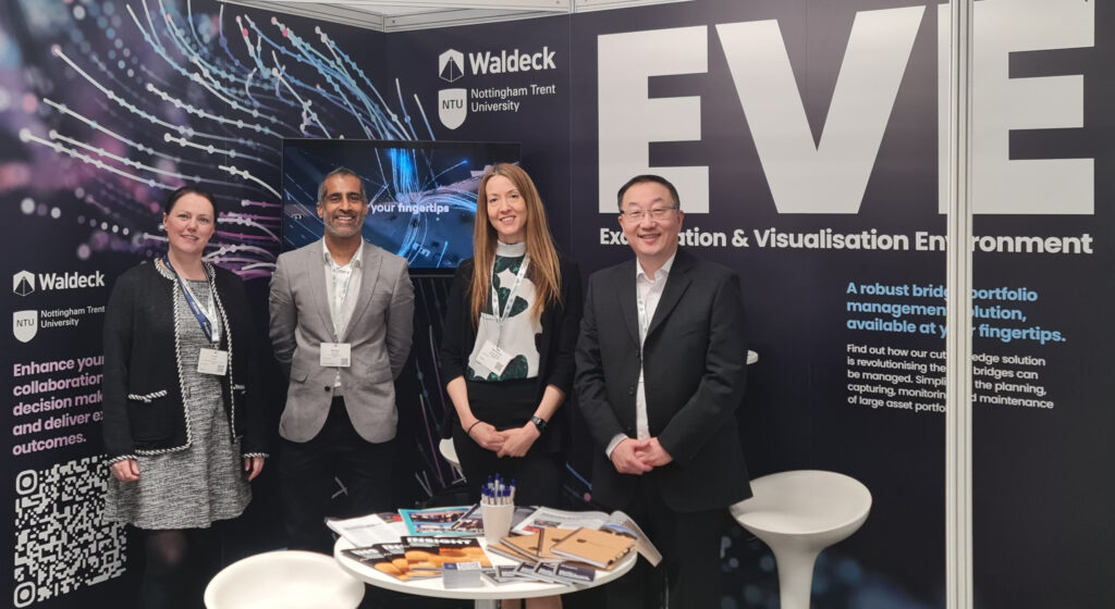 Four people stand smiling at a booth for Waldeck, Nottingham Trent University, and Network Rail, with an "EVE" exhibition banner and promotional materials on the table. The backdrop highlights digital graphics showcasing integration and collaboration.