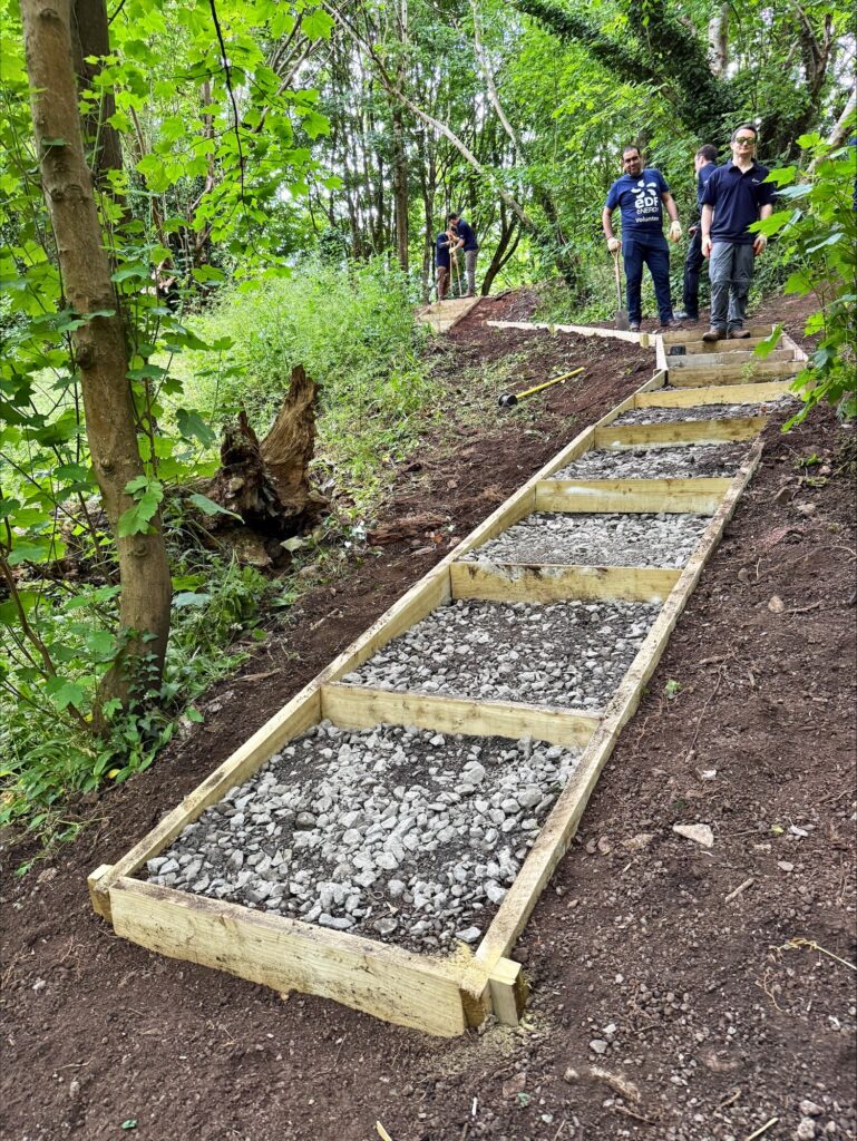 Wooden frames forming steps on a dirt slope are filled with gravel, creating a new pathway in Badock Wood. Several people, part of the Engineering Delivery Team, stand at the top of the slope, appearing to work on the project.