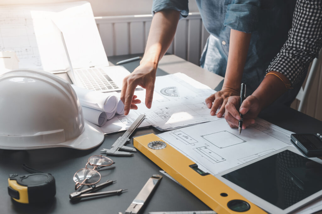 Two people review architectural blueprints on a desk covered with a hard hat, measuring tools, rolled plans, and a tablet—capturing the collaboration between civil engineering and structural engineering in a well-lit workspace.