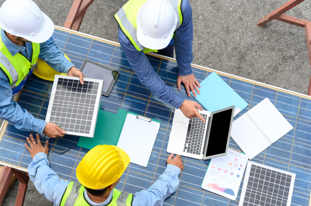 Three construction workers in safety gear examine solar panels, documents, and a laptop on a table covered with solar cells, collaborating on a Power Civils Engineering solar energy project outdoors.