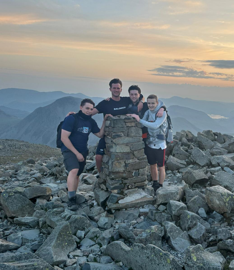 Four young men stand smiling around a stone trig point on a rocky summit at sunset, dressed in hiking gear after completing the National 3 Peaks Challenge, raising funds for St Luke's Sheffield Hospice with stunning mountains in the background.