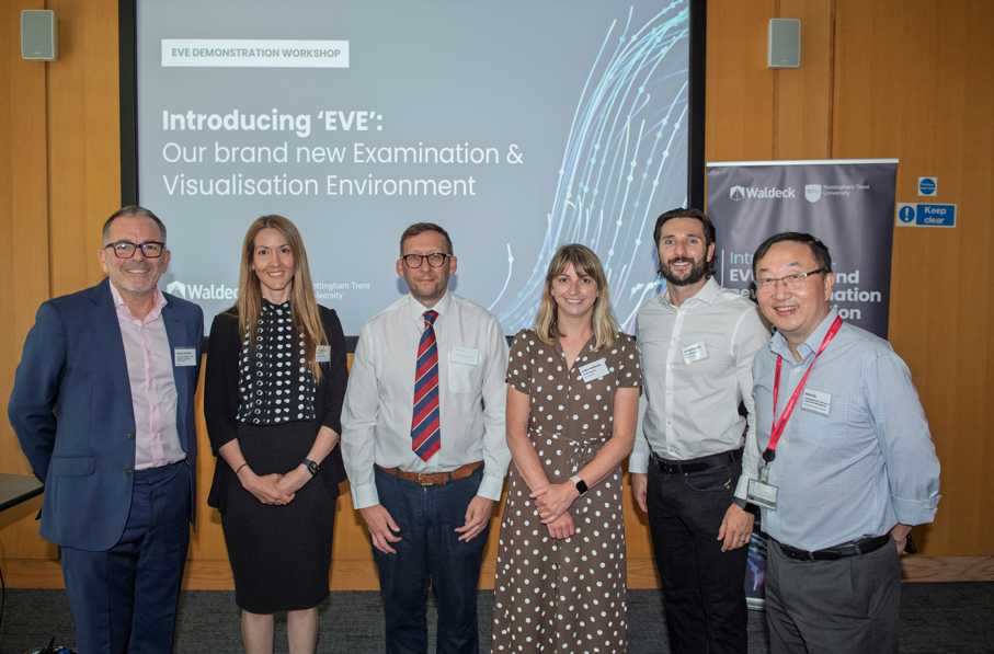 Six people stand in front of a presentation screen displaying "Introducing 'EVE': Our brand new Examination & Visualisation Environment" during an EVE Demonstration workshop at Nottingham Trent University. All are smiling and dressed in business attire.
