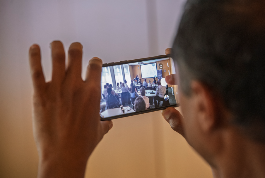 A person holds a smartphone, capturing an EVE Demonstration during a workshop at Nottingham Trent University, where several attendees are seated and listening to a speaker near a screen. The focus is on the phone’s display.