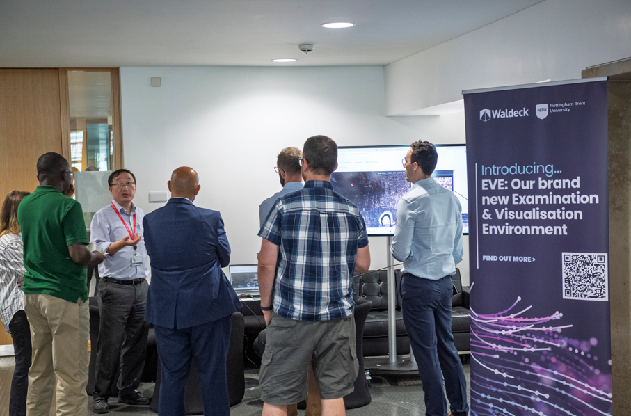 A group of people stand and talk around a screen in an office setting during an EVE Demonstration; a large banner promoting Waldeck’s new Examination & Visualisation Environment, EVE, is visible in the foreground.