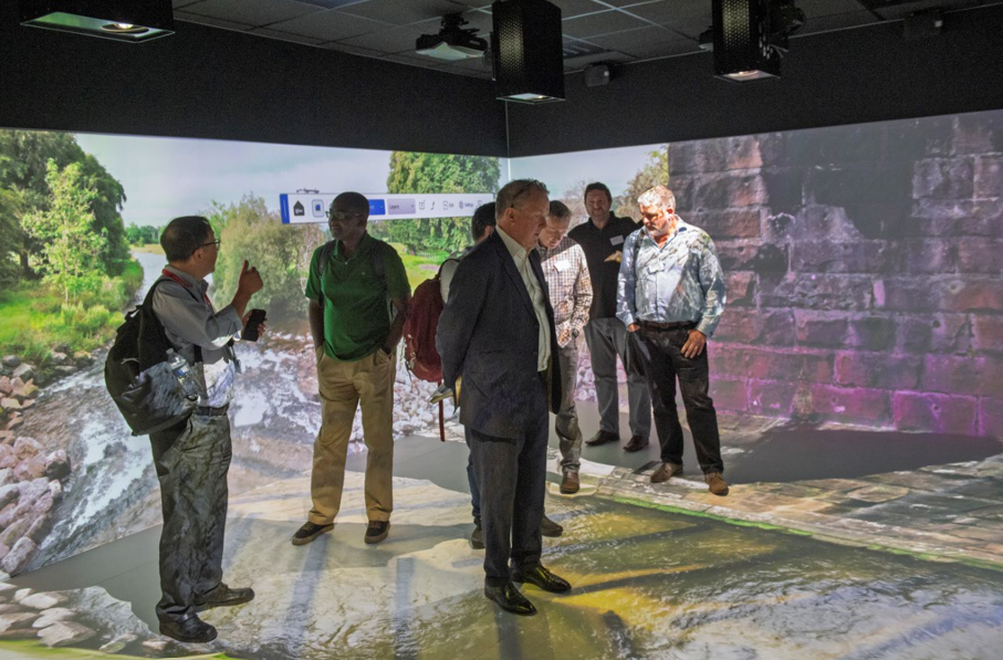 A group of six men stand in a room at Nottingham Trent University with immersive floor and wall projections showing a river scene with trees and a stone structure, likely participating in an EVE Demonstration or Workshop. One man gestures or photographs the group.