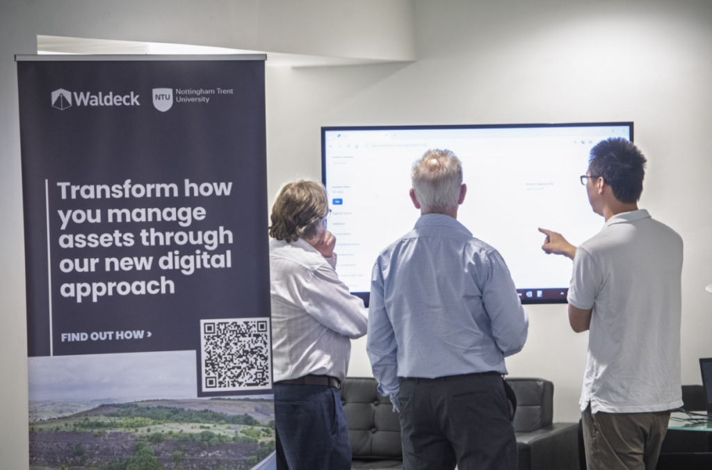 Three men stand in front of a large screen, discussing its content. Next to them is a banner for an EVE Demonstration workshop with a QR code and logos for Waldeck and Nottingham Trent University.